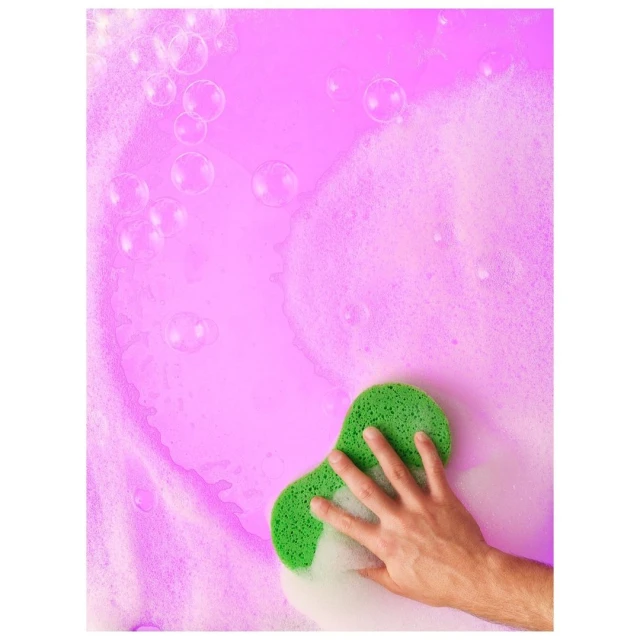 Hand holding green sponge with soap suds on a pink background. Soft bubbles of soapy water and foam surround the hand and sponge.