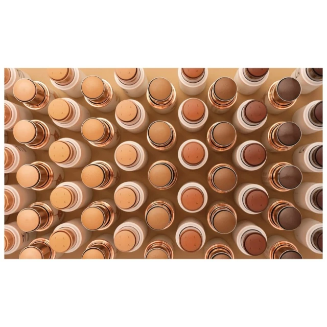 Overhead shot of multiple rows of white and gold cosmetic concealer sticks in varying neutral skin tones, for beauty and makeup concepts.