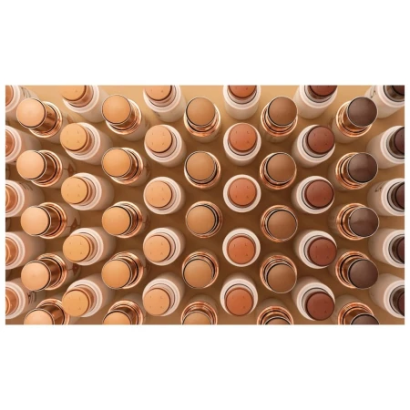 Overhead shot of multiple rows of white and gold cosmetic concealer sticks in varying neutral skin tones, for beauty and makeup concepts.