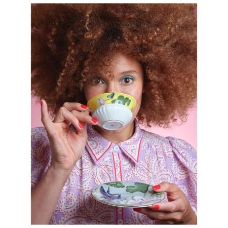 Woman with large auburn afro drinking tea from a floral teacup and saucer in front of a pink background. Her dress is patterned.