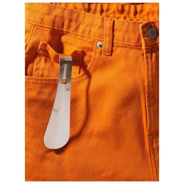 Close-up of orange denim jeans with a silver shoehorn clipped to the front pocket, set against a neutral backdrop. Fashion.