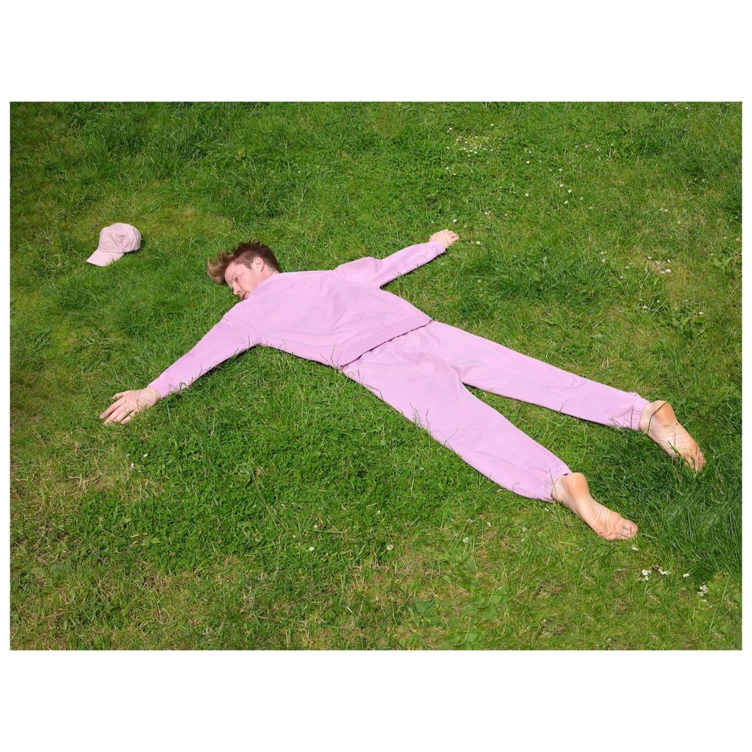 A blond adult male in a pink tracksuit lies barefoot on a green lawn with arms and legs spread. A matching pink cap is nearby.