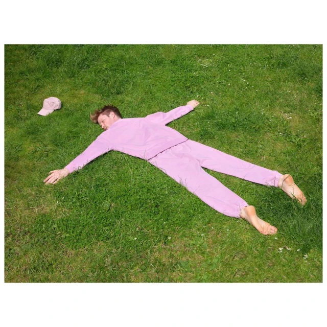 A blond adult male in a pink tracksuit lies barefoot on a green lawn with arms and legs spread. A matching pink cap is nearby.