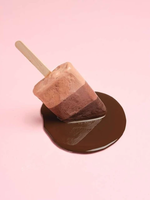 Melting tri-color ice cream popsicle on a stick with chocolate puddle on a light pink background in a studio food shot.
