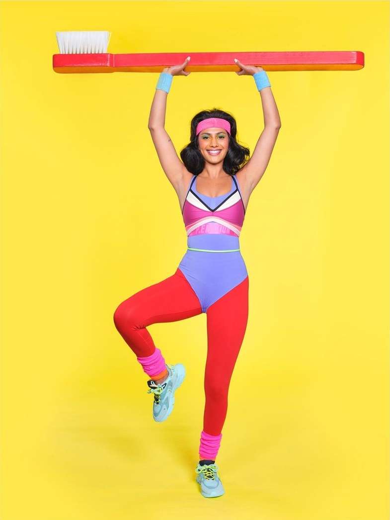 Woman in 80s aerobics outfit holds giant red toothbrush over her head against a yellow background in a full studio shot.