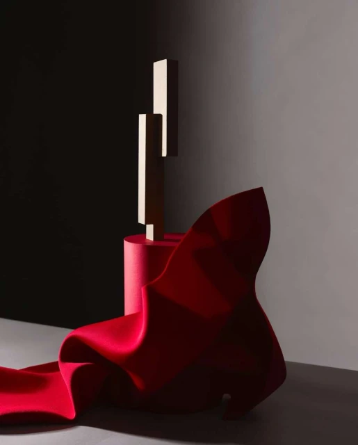 Abstract studio shot of red fabric draped around geometric forms on red cylinder against gradient grey and dark grey backdrop.