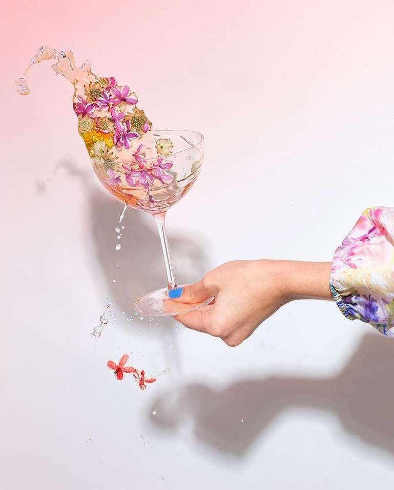 Floral arrangement inside a coupe glass with water spilling out, held by an adult female hand with blue nail polish against a pink backdrop.