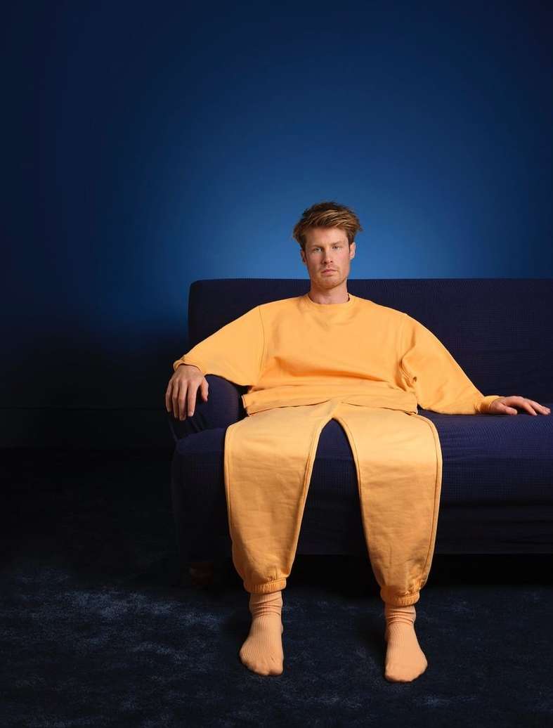 A man in yellow lounge wear sits on a dark blue couch in front of a dark blue background, creating a simple yet striking studio portrait.