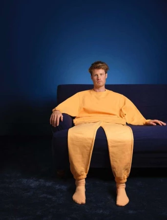 A man in yellow lounge wear sits on a dark blue couch in front of a dark blue background, creating a simple yet striking studio portrait.