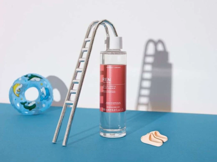 Product shot of Ren Smooth Prep and Plump Essence in clear bottle, next to pool ladder and summer-themed accessories on blue surface.
