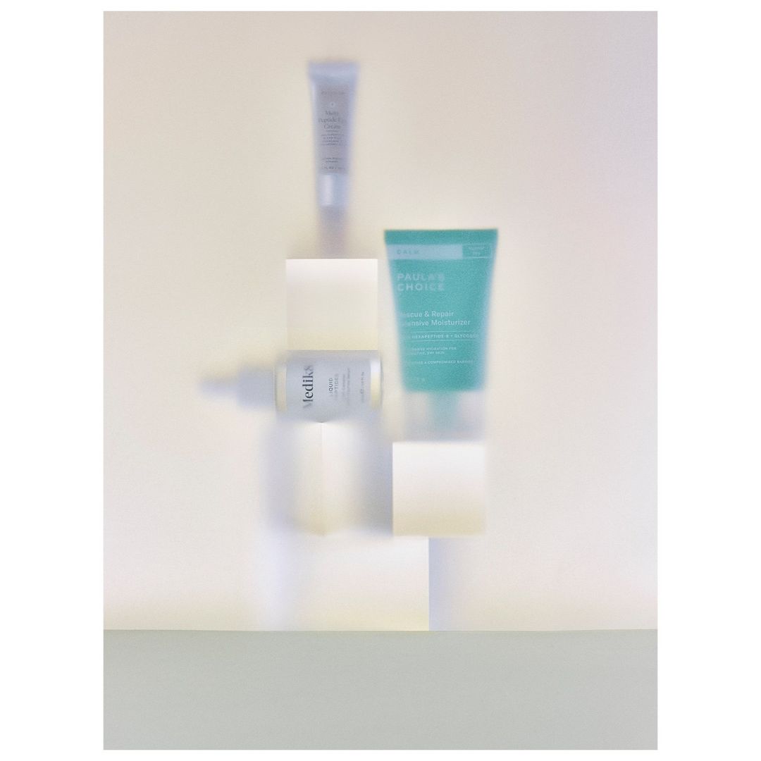 Cosmetic product tubes and bottles arranged on white blocks against a light yellow background in a still life product shot.
