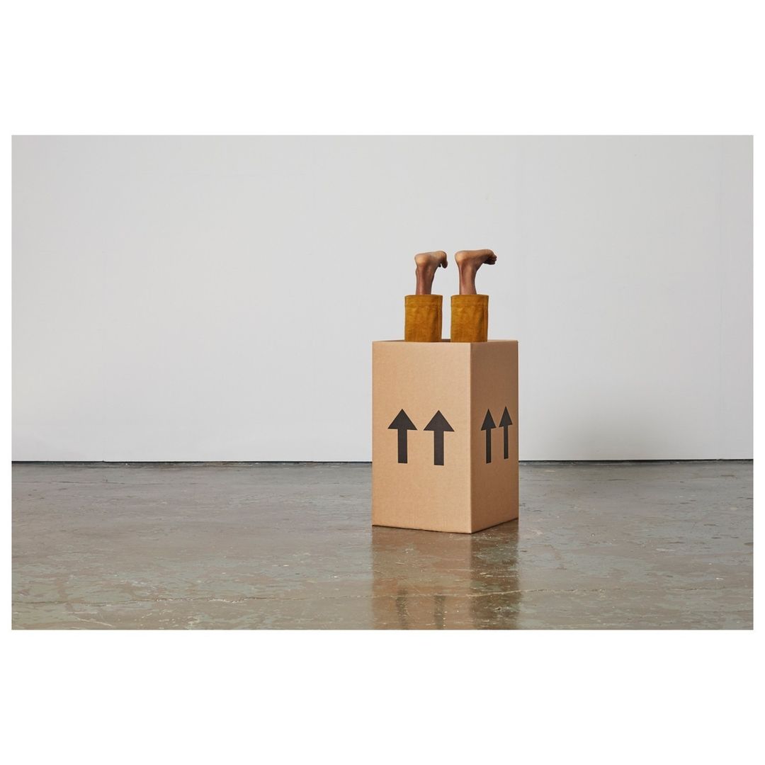 Conceptual still life of mannequin feet sticking out of a cardboard box with black upward arrows on a grey concrete floor against a white wall.