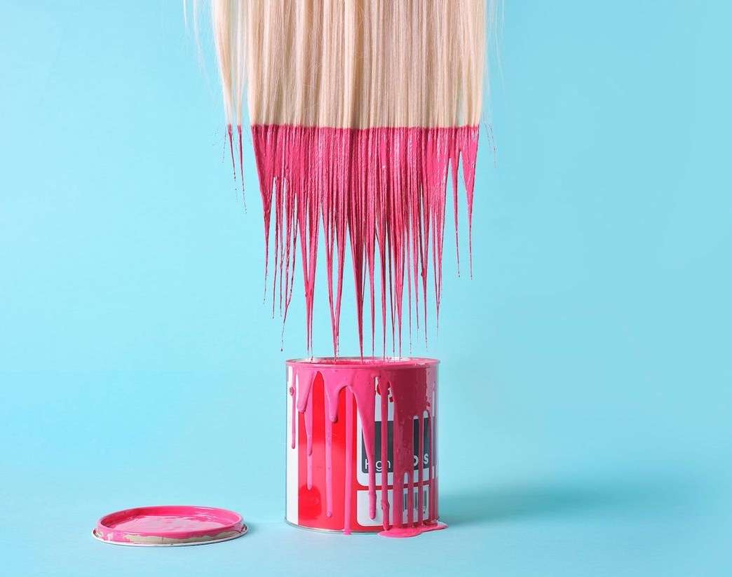 Blonde hair dipped in pink paint above a paint can and lid on a light blue background, creating an abstract beauty theme.