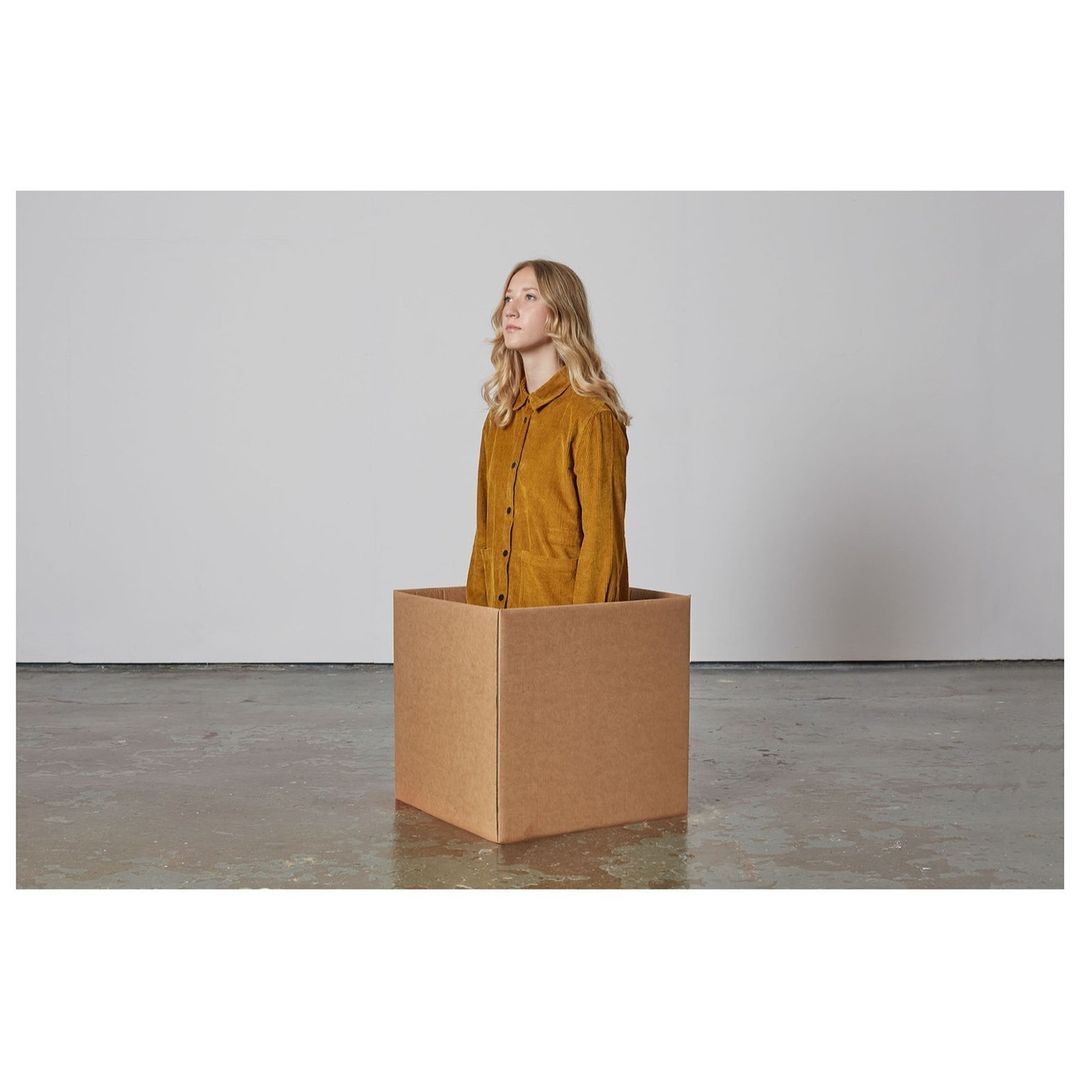 A fair-skinned young woman with blonde hair stands inside a brown cardboard box in a minimalist setting with a concrete floor.