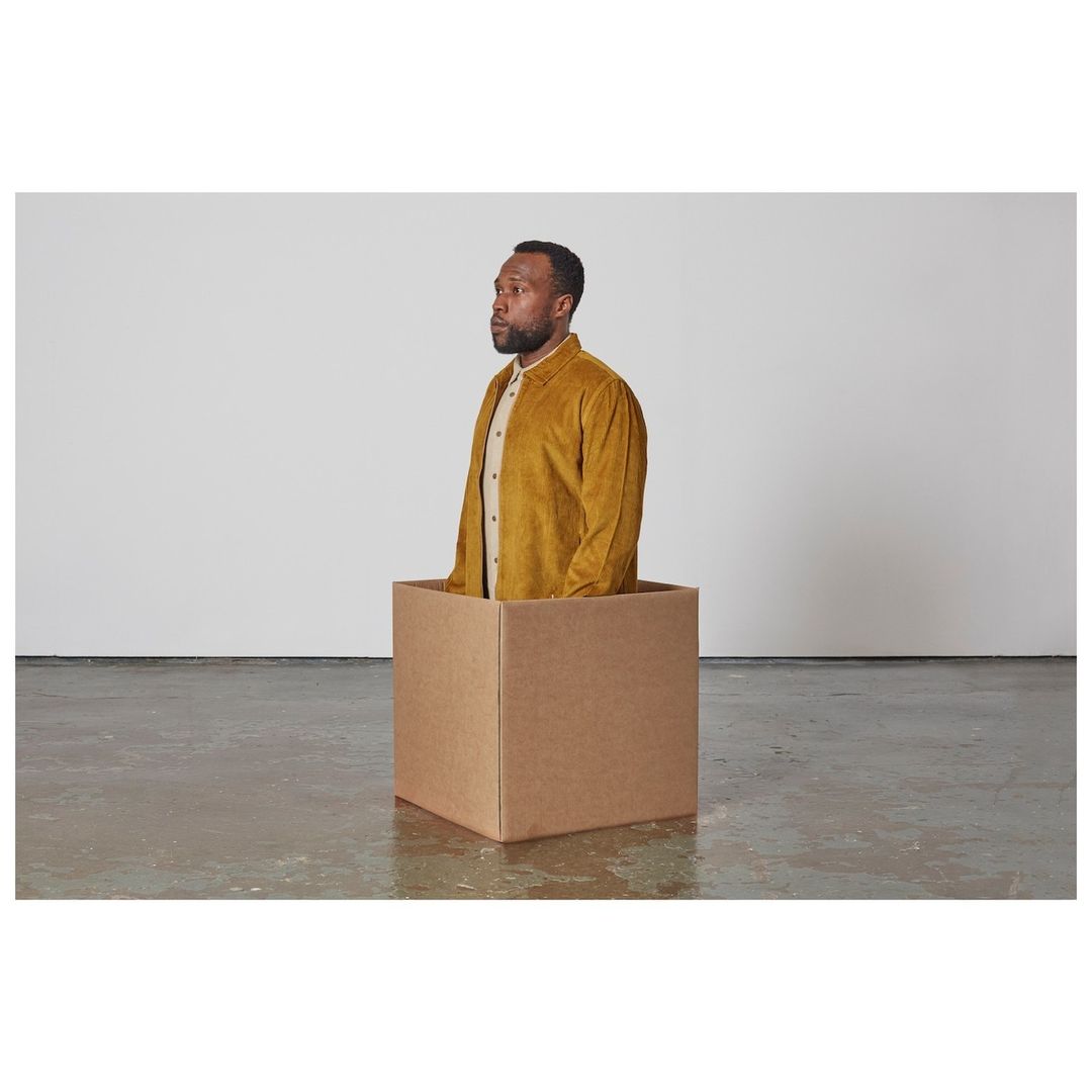 Portrait of an African-American adult male in a yellow jacket inside a brown cardboard box, against a plain white wall and concrete floor.