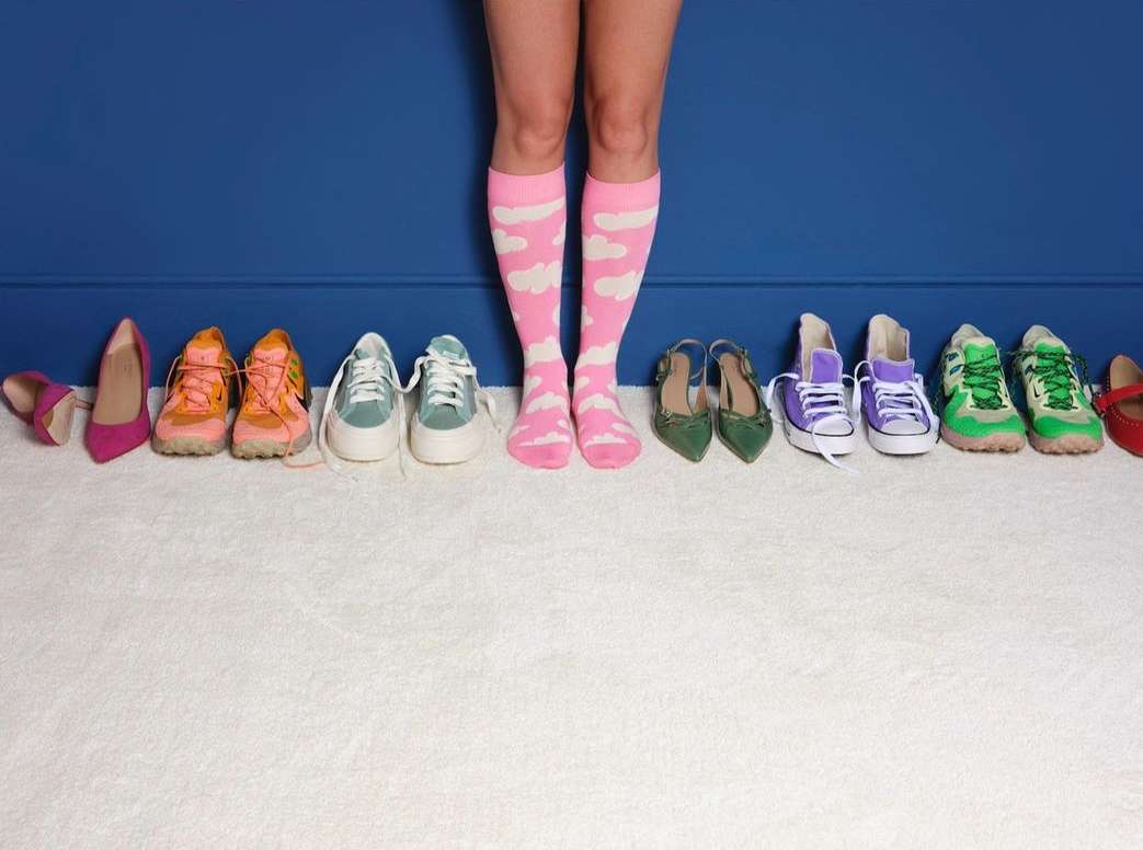Legs of a person wearing pink socks with white clouds standing in front of an assortment of shoes against a dark blue wall and white carpet.