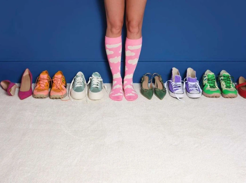 Legs of a person wearing pink socks with white clouds standing in front of an assortment of shoes against a dark blue wall and white carpet.