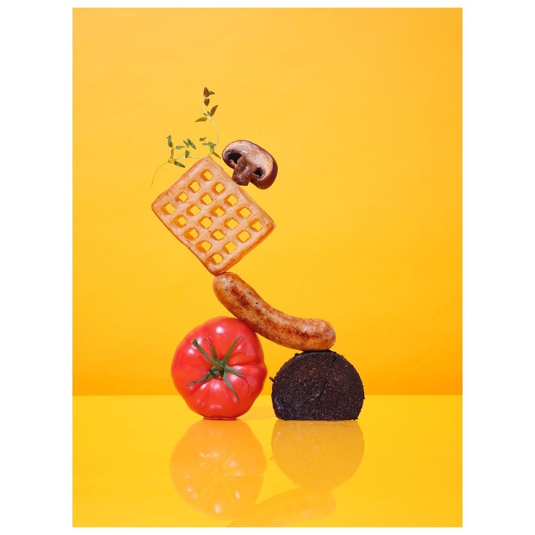 Still life food photograph of a waffle, mushroom, sausage, tomato, and dark ball stacked on a yellow reflective surface.