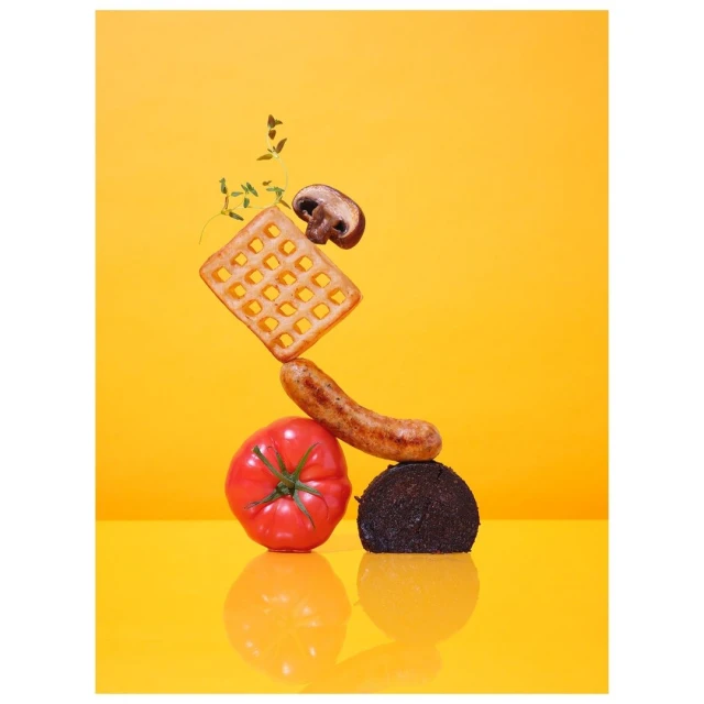 Still life food photograph of a waffle, mushroom, sausage, tomato, and dark ball stacked on a yellow reflective surface.