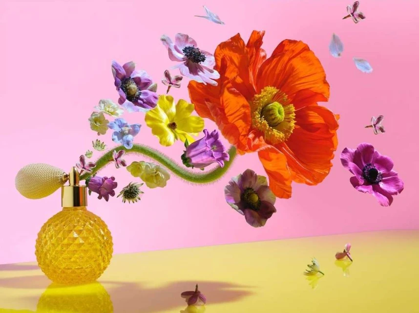 Still life of a yellow faceted perfume bottle emitting a stream of colourful flowers against a pink background, studio lighting.