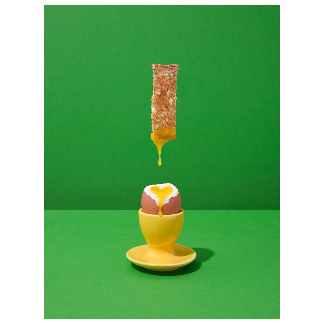 Overhead studio shot of dripping egg yolk on toast above a boiled egg in a yellow egg cup set against a vivid green background.