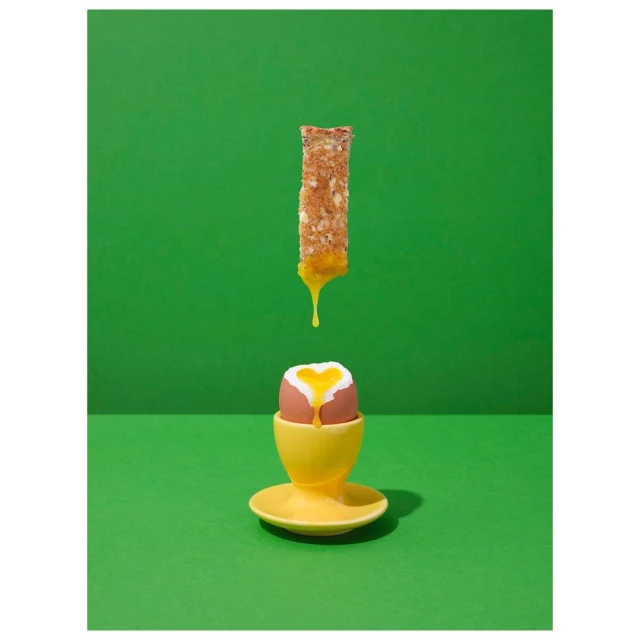 Overhead studio shot of dripping egg yolk on toast above a boiled egg in a yellow egg cup set against a vivid green background.