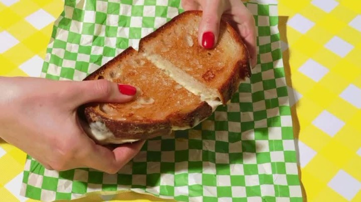 A close-up shot features a person holding a grilled cheese sandwich on a checkered napkin, with bright yellow checkered surface visible.