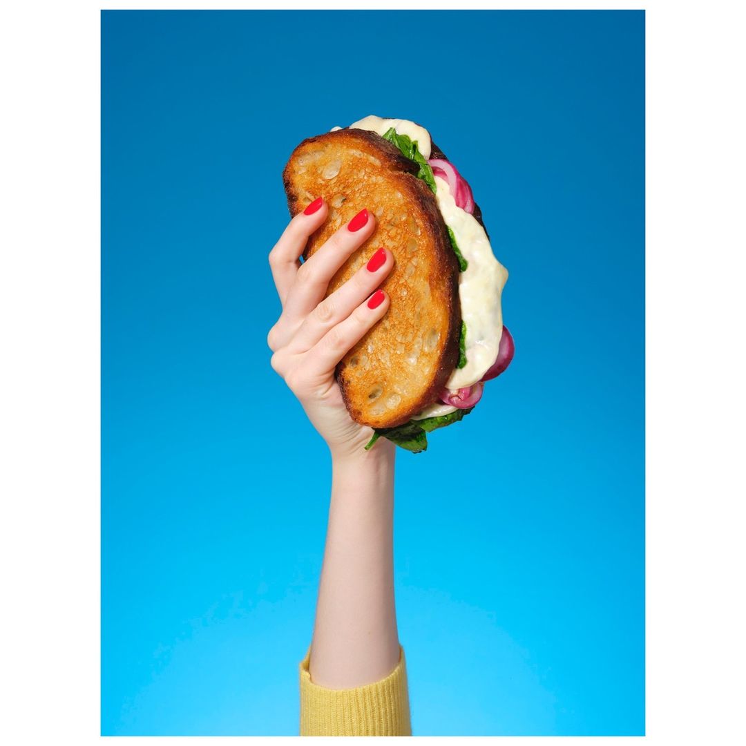 Hand holding a grilled cheese sandwich with lettuce and red onion against a vibrant blue background in a vertical studio shot.