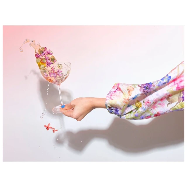 Woman's hand holds cocktail glass overflowing with colorful flowers and water droplets, set against a soft white to pink gradient backdrop.
