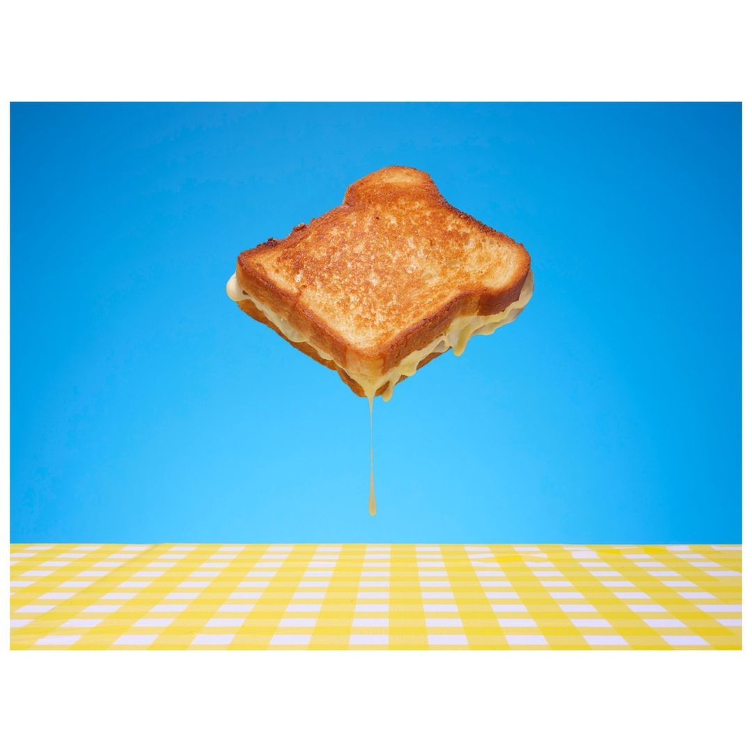 Elevated view of a grilled cheese sandwich with cheese dripping against a blue backdrop and yellow checkered tablecloth. Food and beverage.
