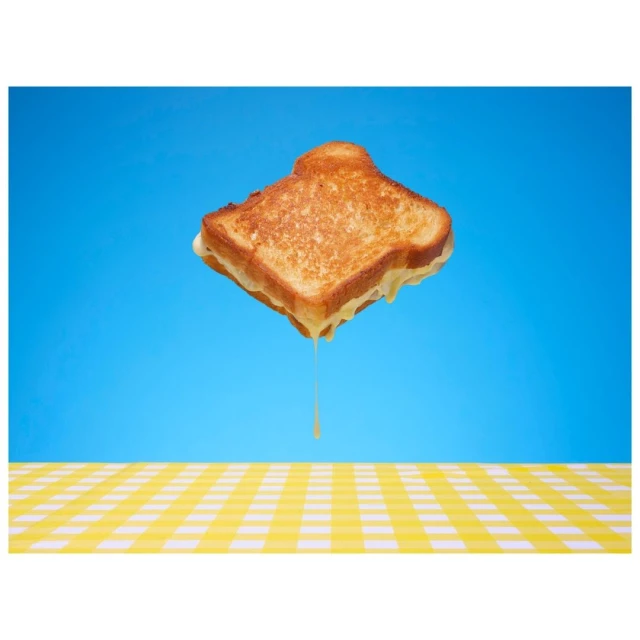 Elevated view of a grilled cheese sandwich with cheese dripping against a blue backdrop and yellow checkered tablecloth. Food and beverage.