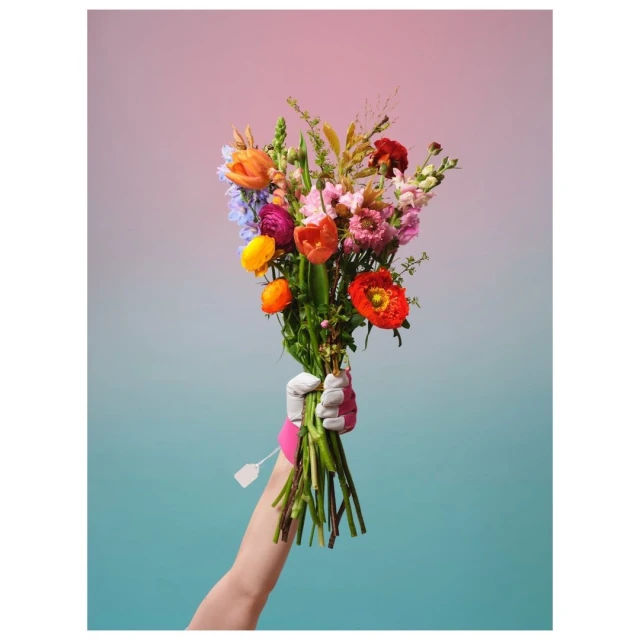 A vibrant floral bouquet featuring tulips, poppies, and other mixed flowers is held by a gloved hand against a soft pink and teal background.