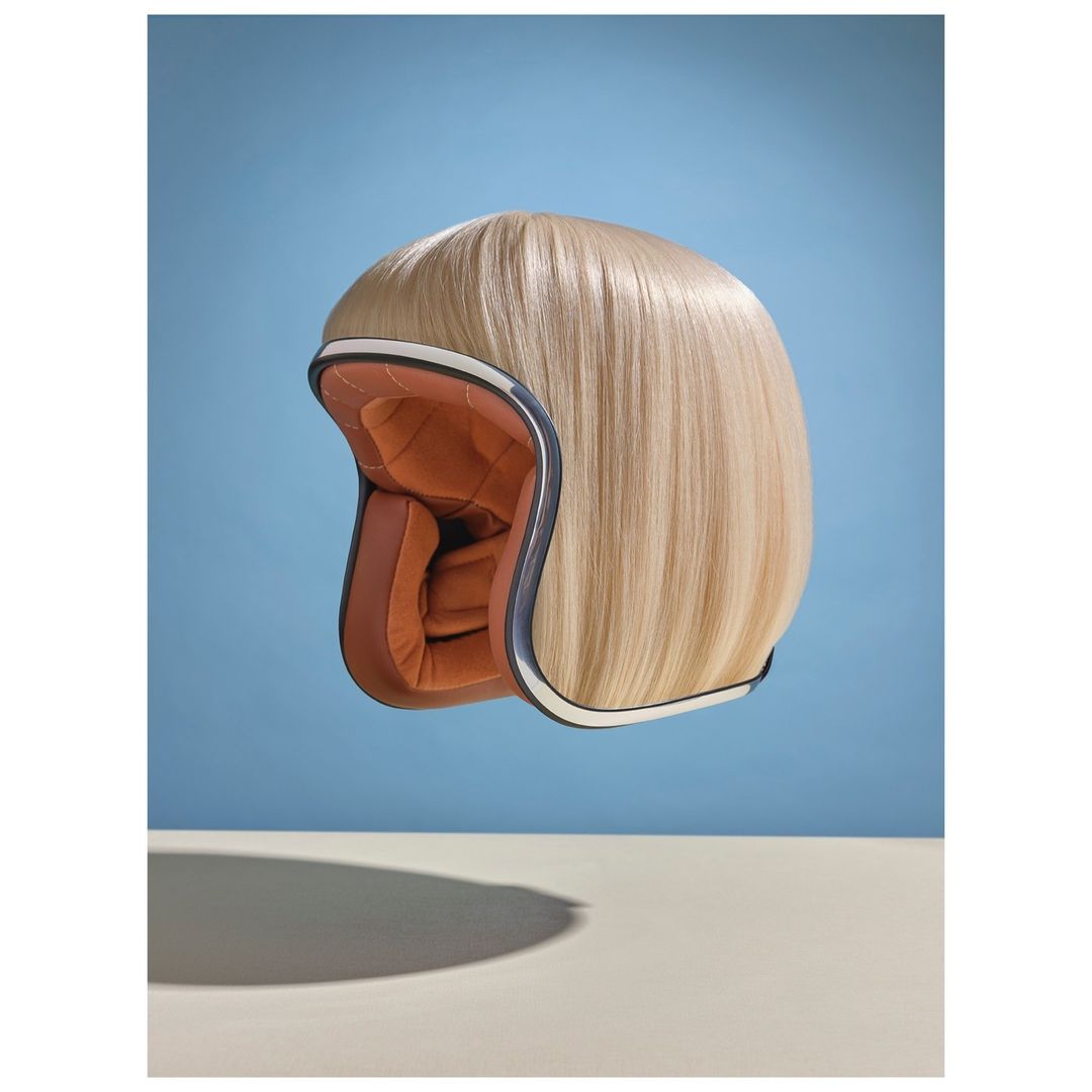 Motorcycle helmet with blond wig on display. Conceptual still life against a blue background featuring retro-style headgear.