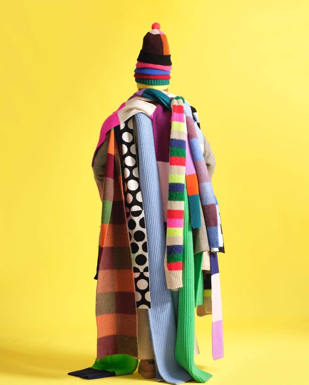 Person from behind wearing multicolored knit hat and multiple scarves against bright yellow backdrop in a studio setting.