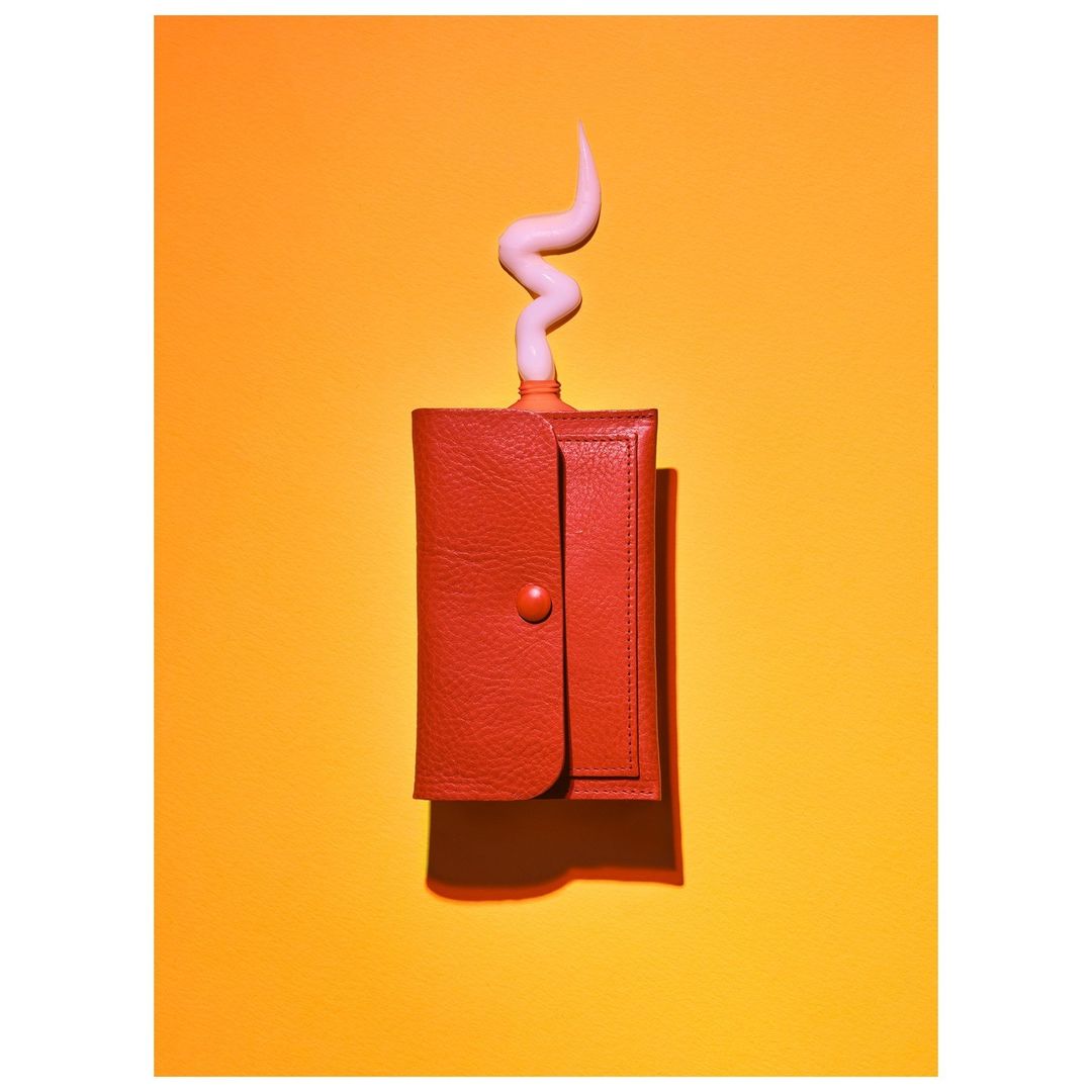 Red wallet with a surreal cream-colored squiggly shape emerging from it, set against a bright orange background. Clean, studio shot.