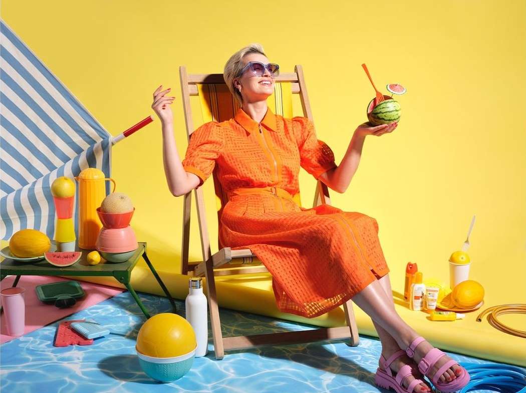 A woman in an orange dress lounges on a wooden chair, holding a watermelon cocktail, against a vibrant yellow and blue studio backdrop.