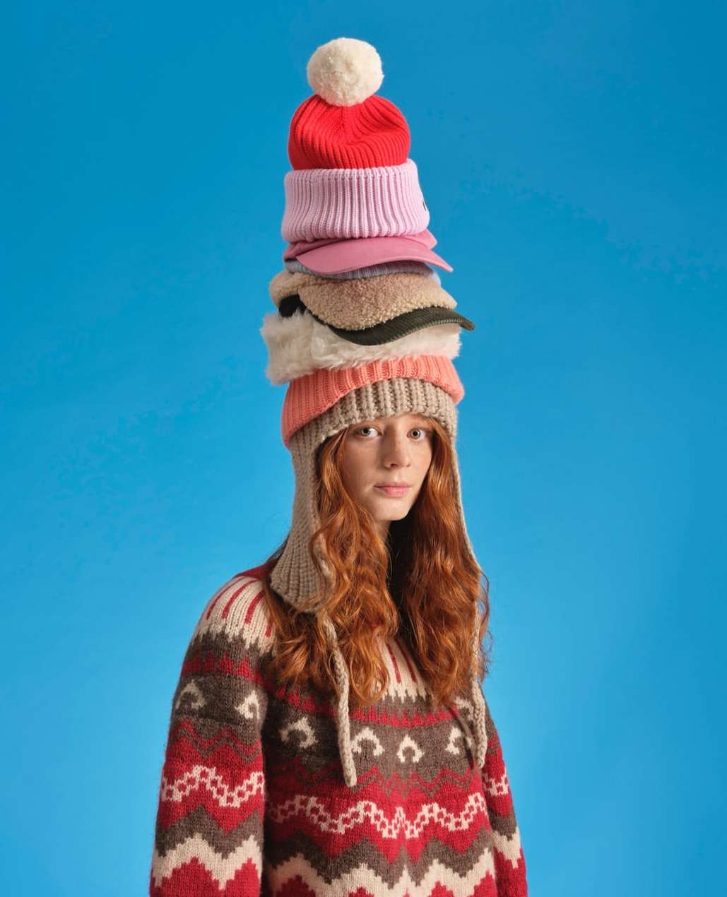 Woman with red hair wearing patterned sweater and a stack of colorful hats on her head against a bright blue studio backdrop.