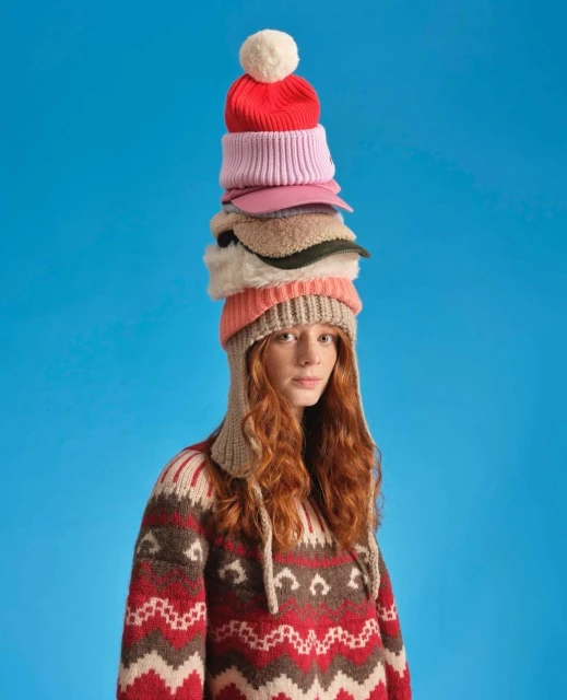 Woman with red hair wearing patterned sweater and a stack of colorful hats on her head against a bright blue studio backdrop.