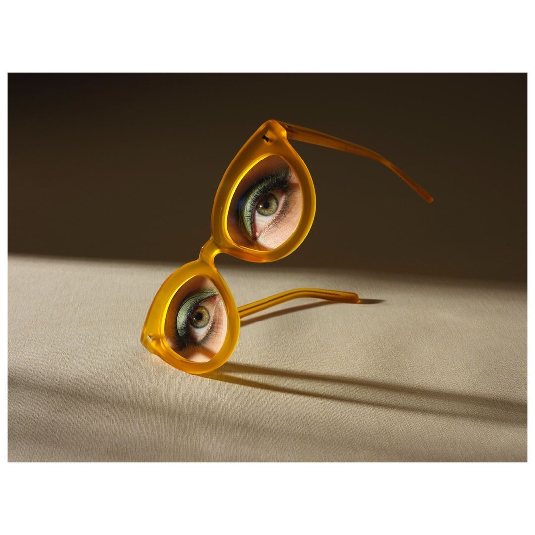 Pair of yellow eyeglasses reflecting a close-up of a woman's eye with green eyeshadow, angled on a beige surface, lit by natural light.