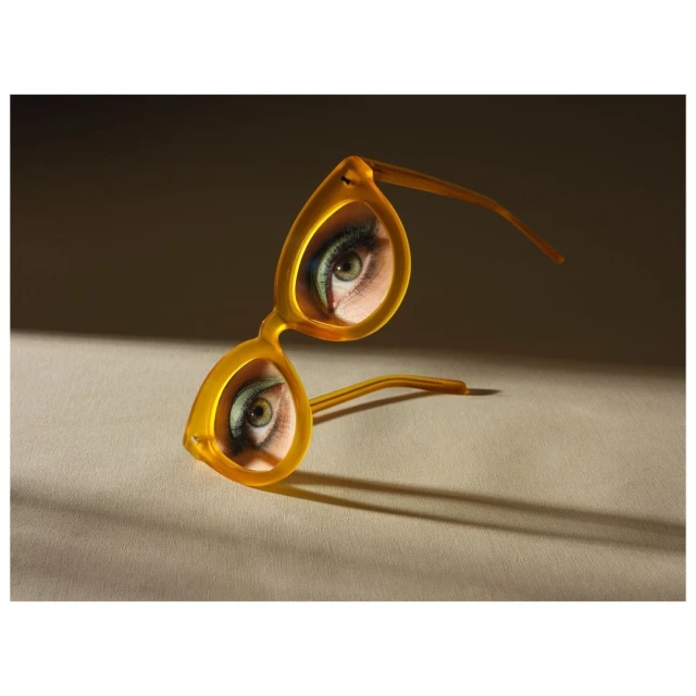 Pair of yellow eyeglasses reflecting a close-up of a woman's eye with green eyeshadow, angled on a beige surface, lit by natural light.