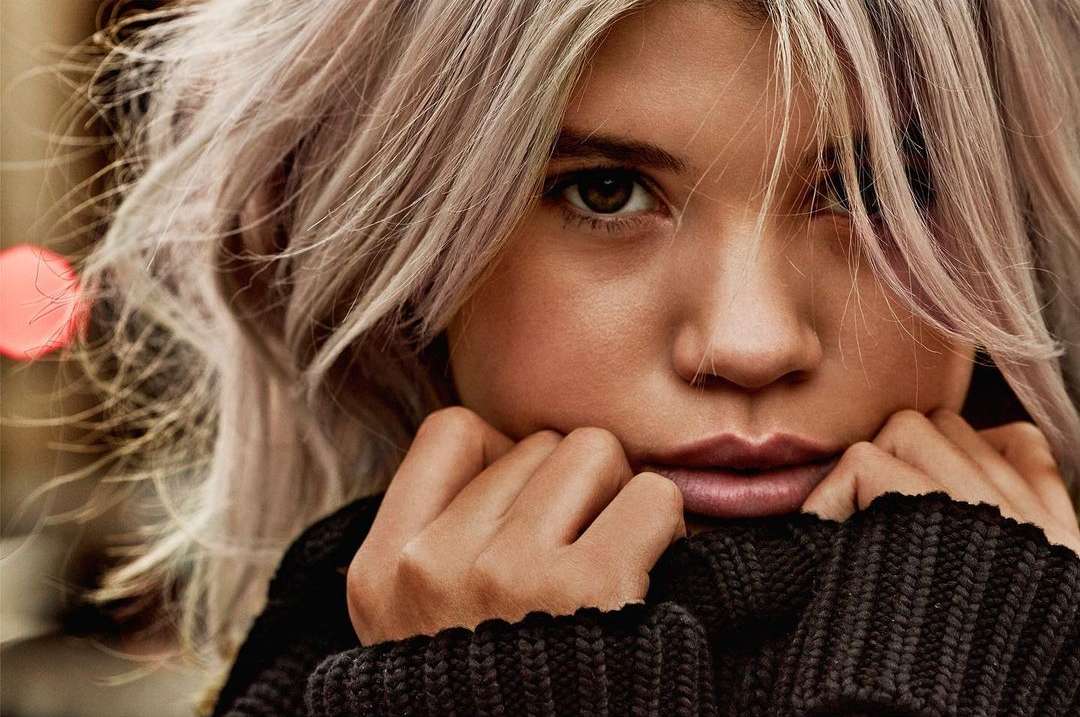 Close-up portrait of a blonde adult female with her hands near her face and wearing a dark knit sweater in soft, diffused lighting.