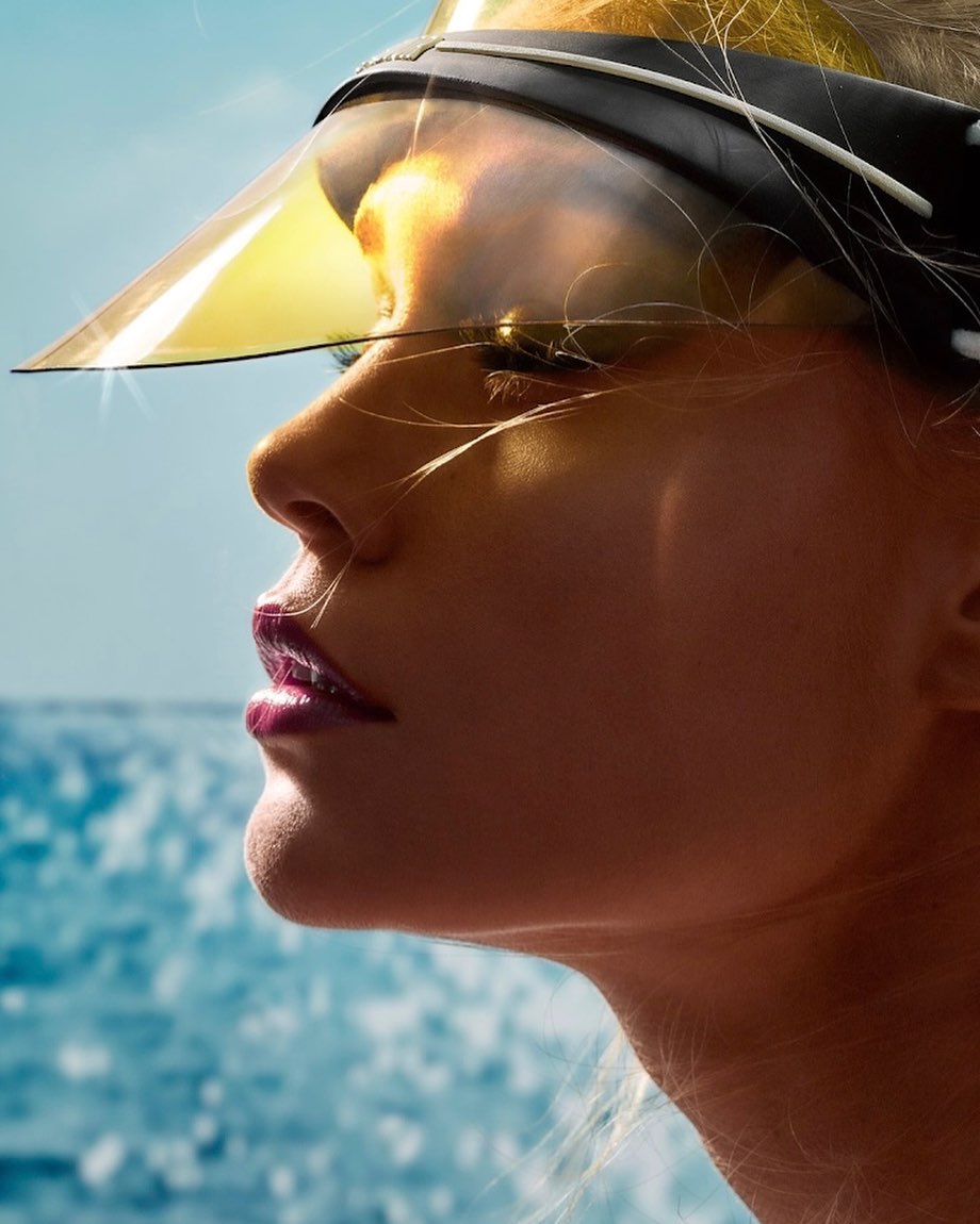 Close-up of a woman's face with blonde hair, glossy lips, and a yellow visor against a bright blue sky and blurred ocean backdrop.