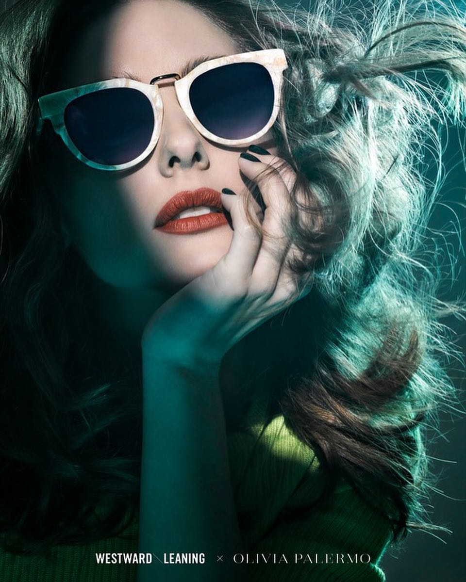 A woman poses indoors wearing cream-colored sunglasses, red lipstick, and a green top, with her hand touching her face.