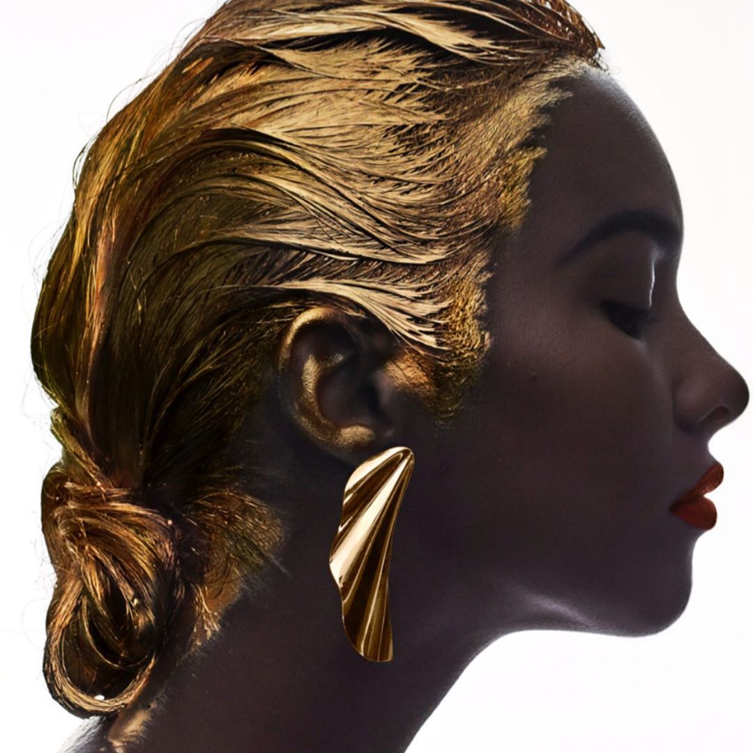 Profile of a dark-skinned woman with golden paint covering her hair and skin. The focus is on her face, gold earring, and red lipstick.