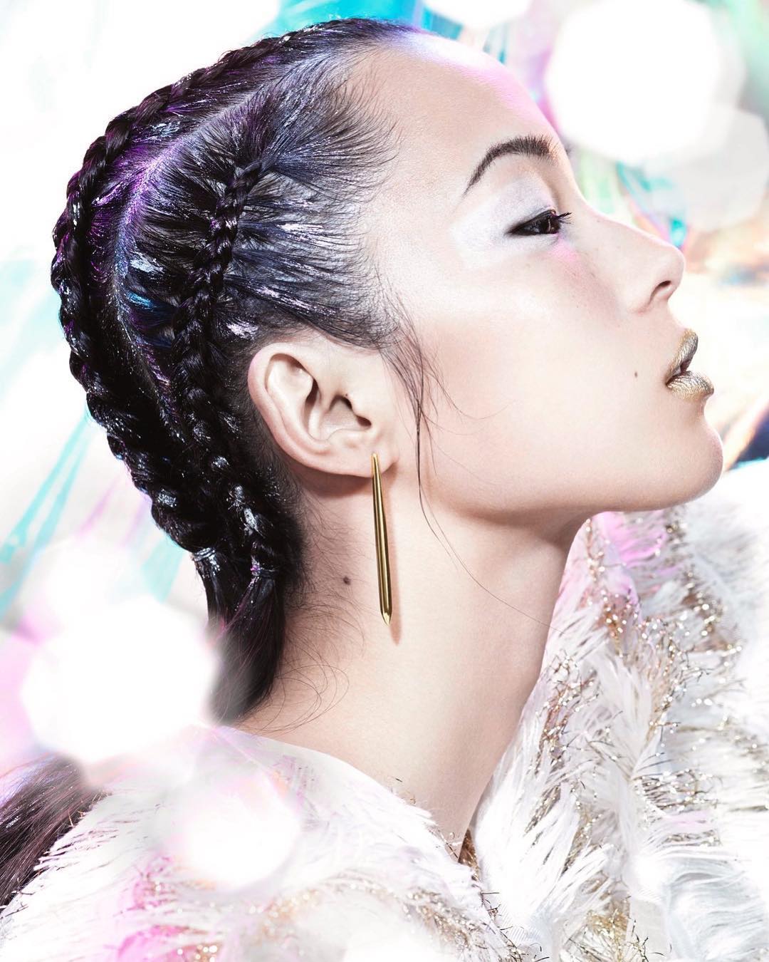 Profile view of woman with braided hair, gold lipstick, and white eyeshadow, wearing long gold earrings, against an out-of-focus background.