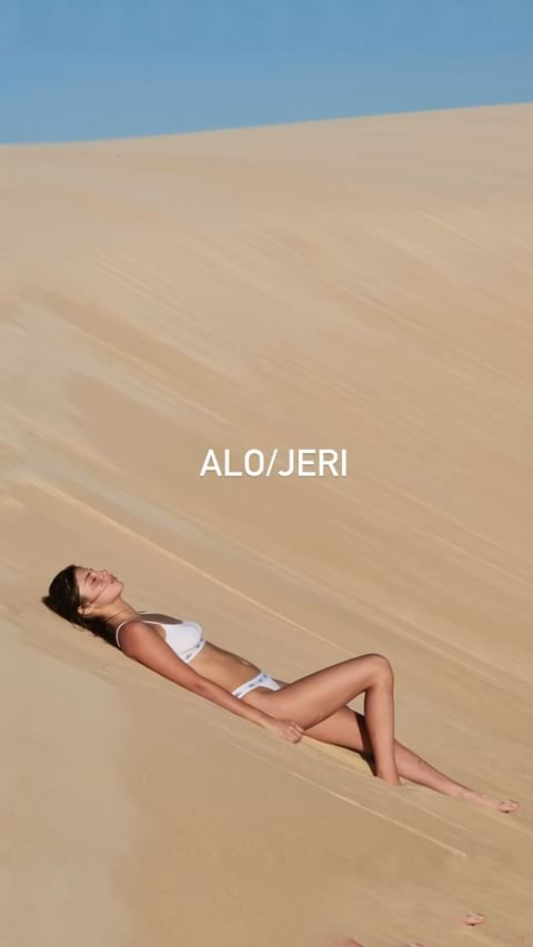 Woman in a white bikini lying on a sand dune against a blue sky, sunbathing on the sand in bright sunlight.