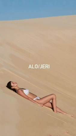 Woman in a white bikini lying on a sand dune against a blue sky, sunbathing on the sand in bright sunlight.