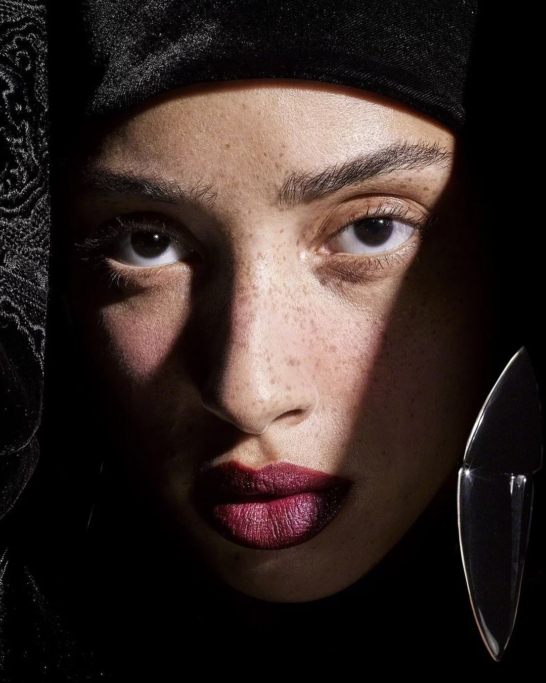 Close-up portrait of a woman with freckles, deep red lipstick, wearing a black headscarf, and a clear earring, against a dark background.