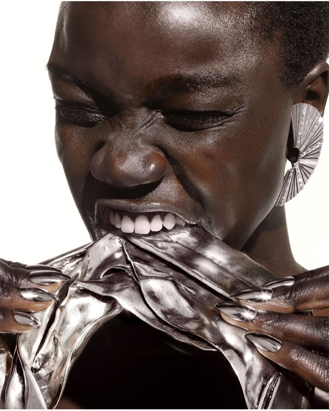 Close-up of a dark-skinned woman with shiny, silver metallic nails biting down on a piece of shiny, silver material.