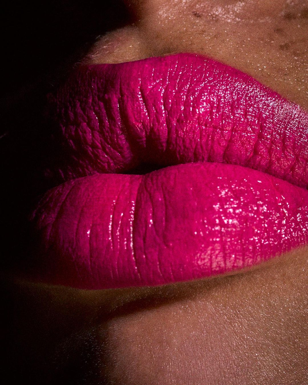 Close-up of vibrant magenta lips with visible skin texture, captured in dramatic lighting that emphasizes the glossy makeup.
